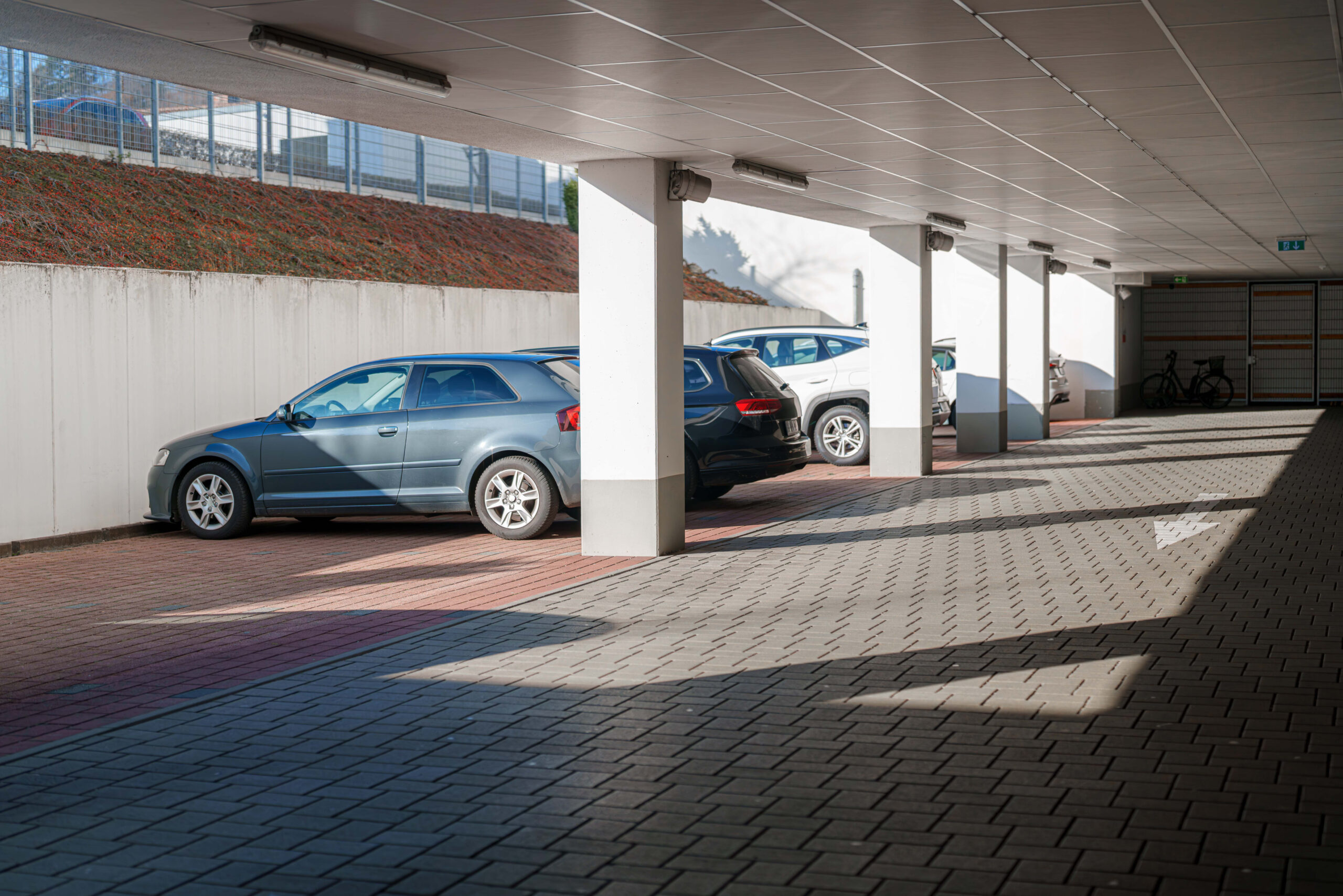 Vehicles are parked in a covered parking lot, with shadows cast by the pillars and roof creating a geometric pattern on the paved ground. A bicycle is visible in the background.